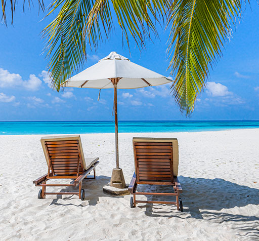 Two lounge chairs under an umbrella on the beach with palm leaves overhead